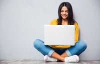 Happy young woman sitting on the floor with crossed legs and using laptop on gray background